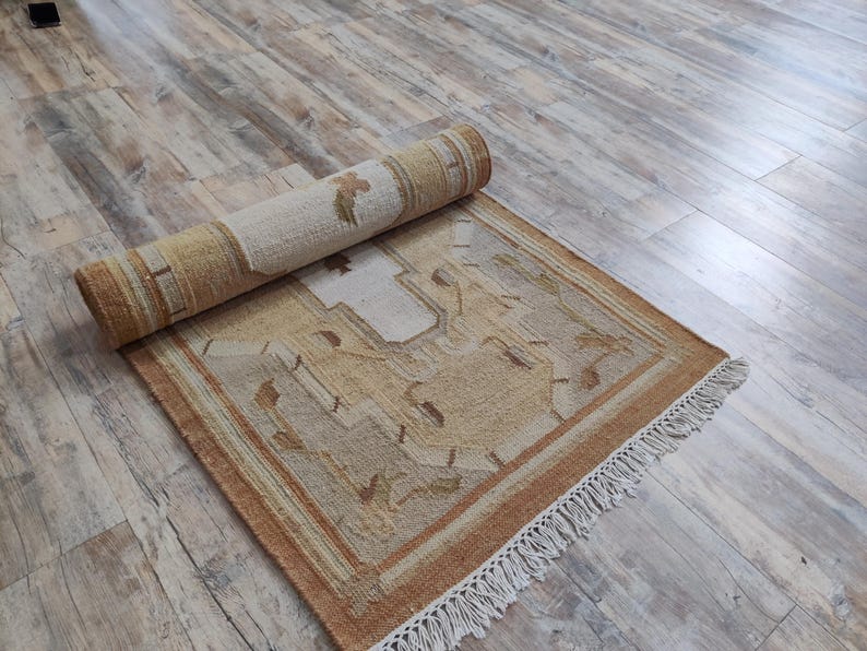 May include: A rolled up beige and brown rug with a geometric pattern and white fringe. The rug is lying on a light wood floor.