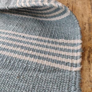 Pu&ograve; includere: Primo piano di un tessuto intrecciato con strisce alternate blu chiaro e bianche. Il tessuto ha un aspetto strutturato e una cucitura visibile lungo il bordo. Il tessuto &egrave; probabilmente un tappeto o un tappetino.