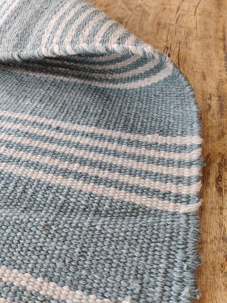 Pu&ograve; includere: Primo piano di un tessuto intrecciato con strisce alternate blu chiaro e bianche. Il tessuto ha un aspetto strutturato e una cucitura visibile lungo il bordo. Il tessuto &egrave; probabilmente un tappeto o un tappetino.
