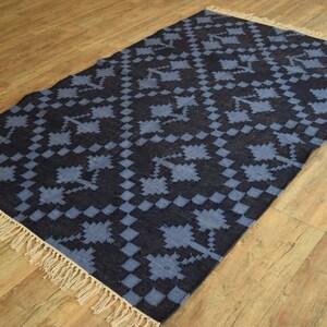 Puede incluir: Una alfombra azul y negra con estampado y borde de flecos. La alfombra tiene un dise&ntilde;o geom&eacute;trico con un patr&oacute;n de cuadros.