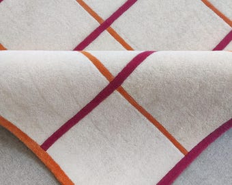 Op de afbeelding: Een wit tapijt met een geometrisch patroon van oranje en roze strepen. Het tapijt is dubbelgevouwen, waardoor de onderkant van het tapijt zichtbaar is.