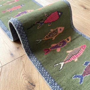 Könnte beinhalten: Ein langer, olivgrüner Läufer mit einem sich wiederholenden Muster aus stilisierten Fischen in verschiedenen Farben wie Pink, Lila und Orange. Der Teppich hat eine strukturierte Bordüre mit blau-weißem geometrischem Muster und den Text HandiRugDecor.