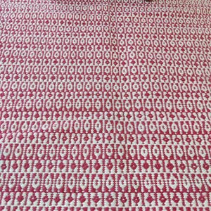May include: A red and white patterned rug with a geometric design. The rug has a repeating pattern of diamond and oval shapes. The rug is made of a woven material and has a textured appearance. A wooden table leg is visible in the upper portion of the image.