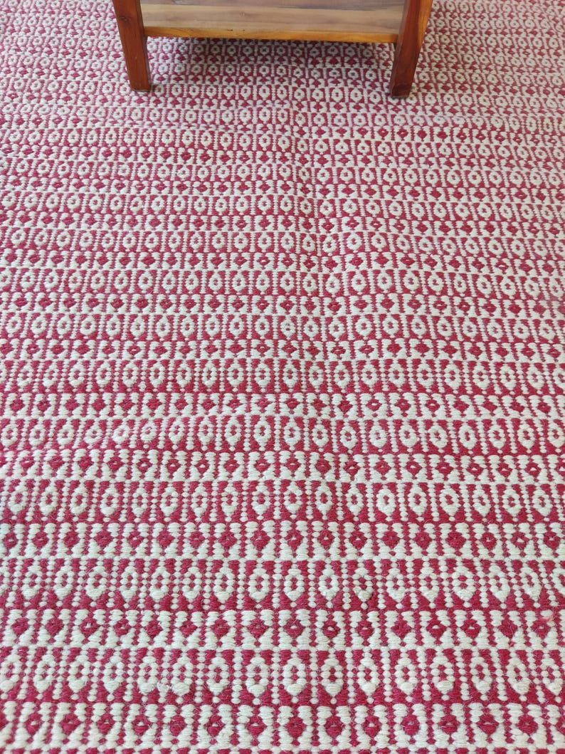 May include: A red and white patterned rug with a geometric design. The rug has a repeating pattern of diamond and oval shapes. The rug is made of a woven material and has a textured appearance. A wooden table leg is visible in the upper portion of the image.