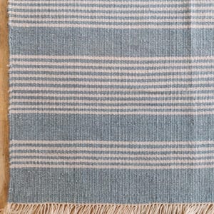 Pu&ograve; includere: Tappeto rettangolare con strisce orizzontali in azzurro e bianco sporco. Il tappeto ha un bordo con frange di colore naturale. La trama tessuta &egrave; visibile, suggerendo una qualit&agrave; artigianale. Adatto per interni.