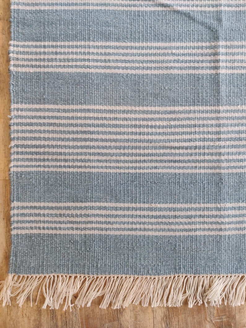 Pu&ograve; includere: Tappeto rettangolare con strisce orizzontali in azzurro e bianco sporco. Il tappeto ha un bordo con frange di colore naturale. La trama tessuta &egrave; visibile, suggerendo una qualit&agrave; artigianale. Adatto per interni.