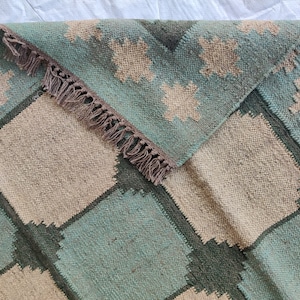 May include: A woven rug with a geometric pattern in shades of green, brown, and beige. The rug has a fringe border.