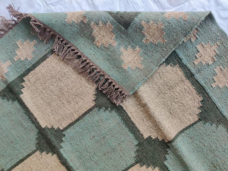 May include: A woven rug with a geometric pattern in shades of green, brown, and beige. The rug has a fringe border.