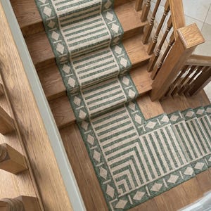 May include: A green and beige patterned stair runner. The runner has a striped design with diamond shapes along the edges. It is laid on wooden stairs. The stairs have a wooden handrail.