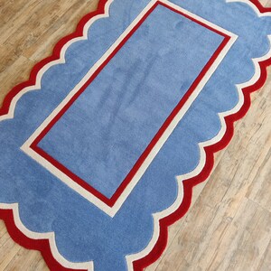May include: A rectangular rug with a scalloped edge. The rug features a light blue center with a red border and a white inner border. The outer edge is red with a white trim, creating a playful and decorative design.