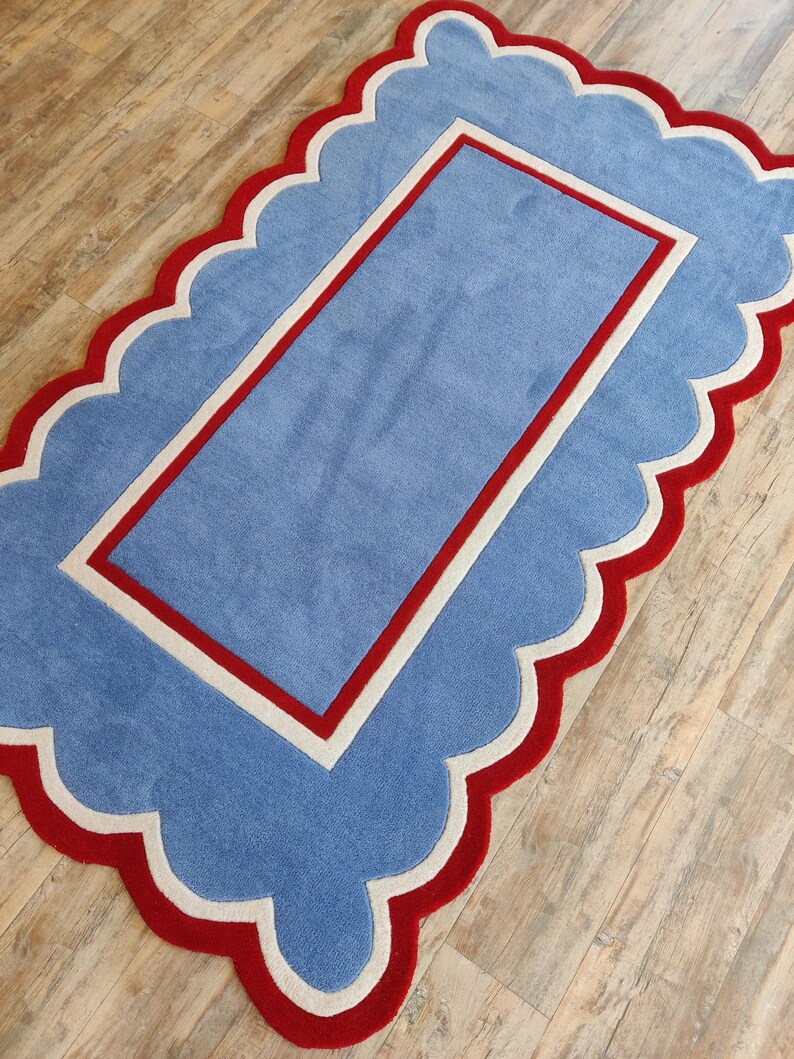 May include: A rectangular rug with a scalloped edge. The rug features a light blue center with a red border and a white inner border. The outer edge is red with a white trim, creating a playful and decorative design.