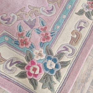 May include: A plush area rug featuring a soft pink center field with a cream-colored border. The design includes carved floral motifs in shades of rose, blue, and sage green, accented by geometric scrollwork patterns along the edges.