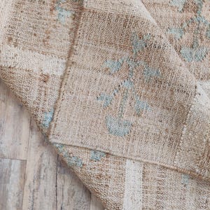 Puede incluir: Alfombra vintage beige y marrón con aspecto desgastado. La alfombra presenta un diseño de patchwork con detalles florales en azul claro. La alfombra está doblada, revelando la parte inferior, y está sobre un suelo de madera.