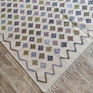 May include: A white rug with a geometric pattern of small squares in shades of blue, green, and brown. The rug has a fringe border.
