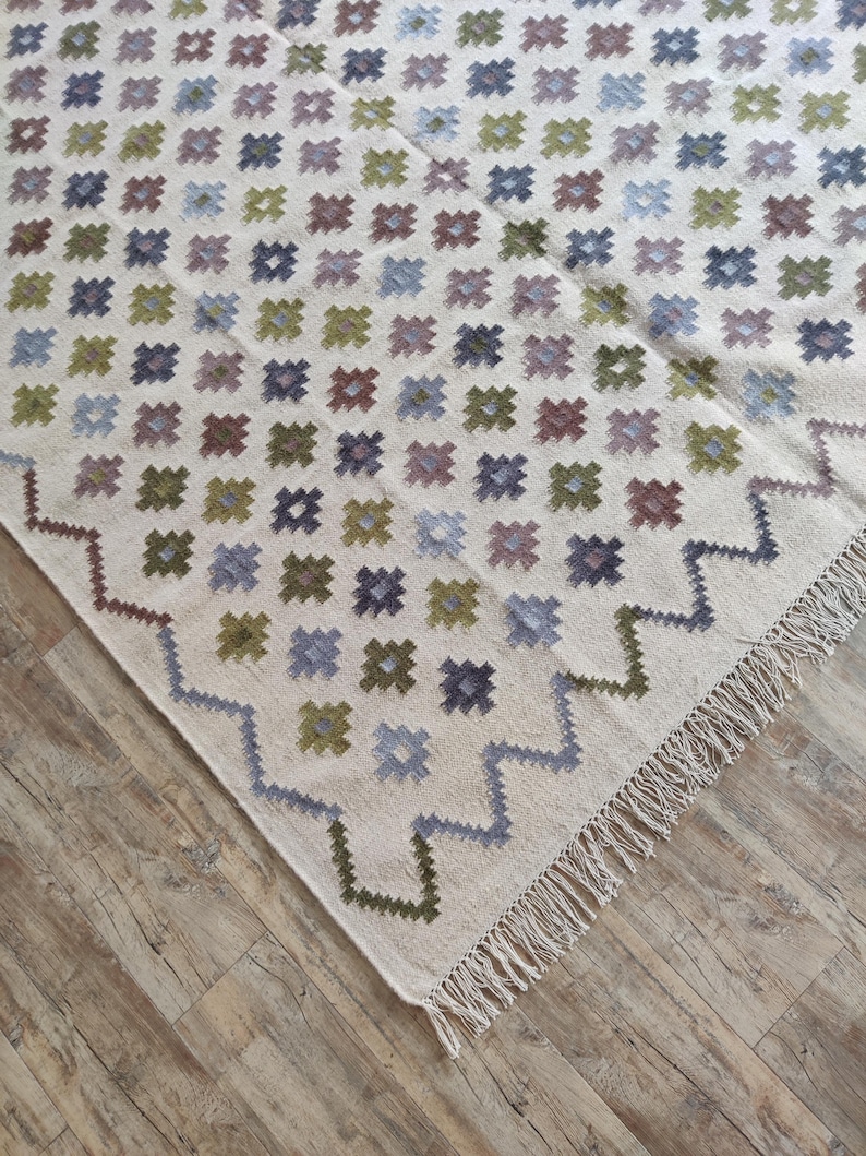 May include: A white rug with a geometric pattern of small squares in shades of blue, green, and brown. The rug has a fringe border.