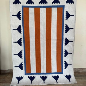 May include: A white rug with a brown and blue geometric pattern. The rug has a border of blue and white flowers.