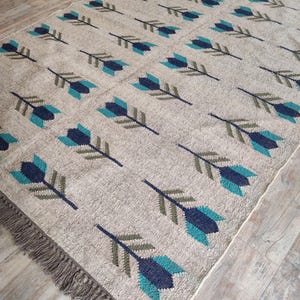 May include: A beige rug with a repeating pattern of teal and dark blue arrows. The rug has a fringe border.