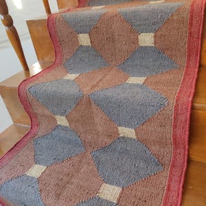 May include: A red, beige, and gray patterned rug with a geometric design. The rug is made of woven fibers and has a fringed edge.