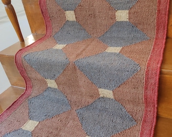 Puede incluir: Una alfombra de diseño geométrico con patrones rojos, beige y grises. La alfombra está hecha de fibras tejidas y tiene un borde con flecos.