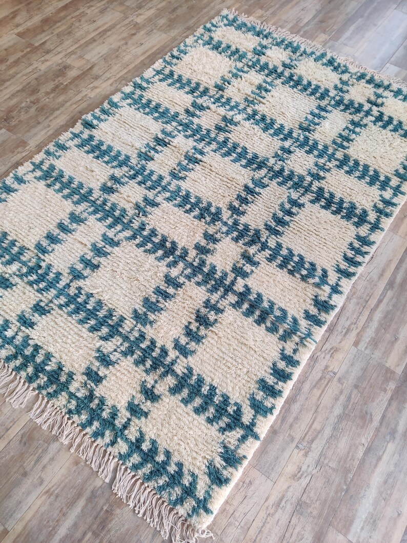 May include: A white and teal blue patterned rug with a fringe border. The rug has a geometric design with a checkered pattern.