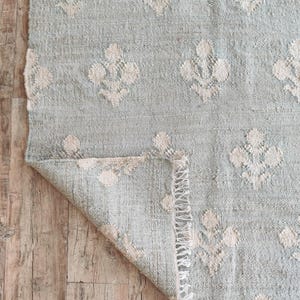 Puede incluir: Una alfombra azul claro con un patrón floral blanco. La alfombra tiene un borde de flecos y está doblada por la mitad.