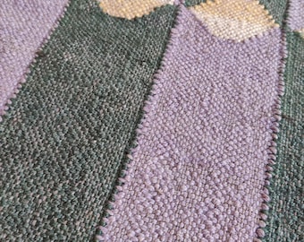 May include: Close-up of a woven textile with a striped pattern. Alternating stripes of muted green and lavender-purple are visible. Beige diamond shapes are stitched into the fabric, adding a decorative element. The texture appears soft and slightly raised.