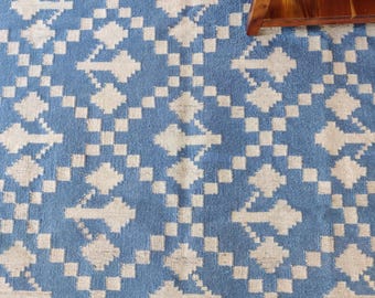 Op de afbeelding: Een blauw en crèmekleurig vloerkleed met een herhalend geometrisch ruitpatroon met gestileerde bloemmotieven in het midden van elke ruitvorm. Het kleed heeft een getextureerd, geweven uiterlijk met een consistent kleurenschema.