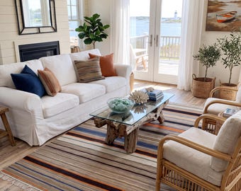 Puede incluir: Una sala de estar con un sofá blanco con cojines azul marino y color óxido, una mesa de centro con tapa de cristal y base de madera flotante, y una alfombra grande con rayas horizontales en tonos marrón, azul y naranja, con flecos.