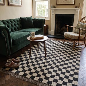 May include: A living room scene featuring a dark green velvet sofa, a round wooden side table, and a black and white checkered rug. The rug has a fringed edge. A wooden chair and a fireplace are also visible.