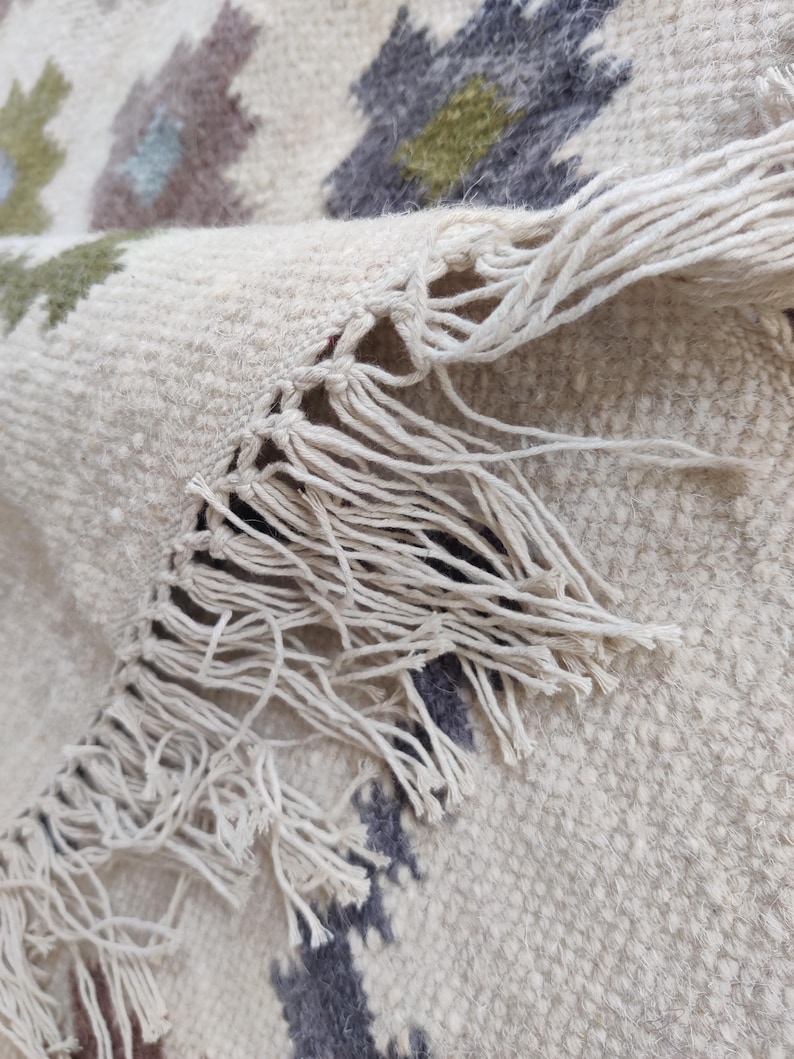 May include: Close-up of a white rug with a fringe border. The rug has a geometric pattern in shades of gray, green, and brown.