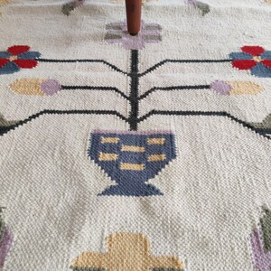 handwoven wool rug features a floral tribal kilim design with soft beige, red, blue, and pastel tones. Made from premium natural wool, this flatweave rug is durable, eco-friendly, and perfect for living rooms, bedrooms, or boho home décor. Each rug is handcrafted by skilled artisans, making it a unique and timeless piece for modern and traditional interiors.