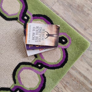 May include: A section of a green and beige rug with a repeating pattern of purple, black, and beige circles. The rug has a soft, textured appearance. A stack of books with the title "How to Use Your Healing Power" rests on the rug.
