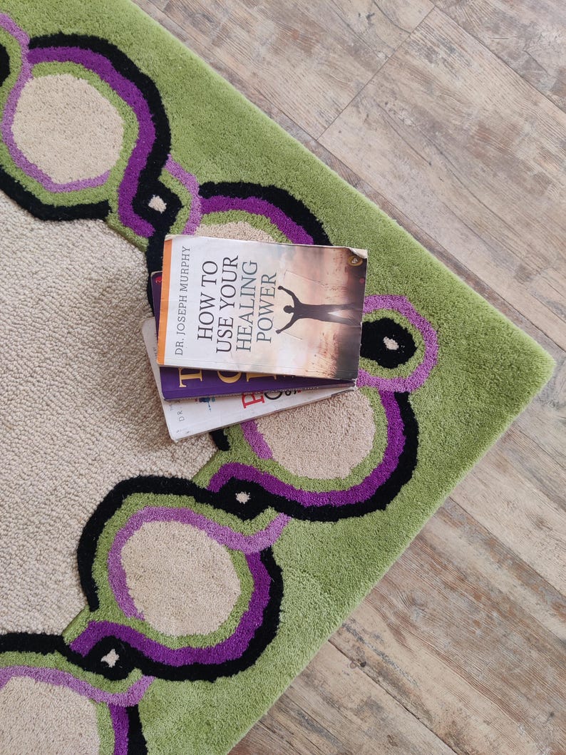 May include: A section of a green and beige rug with a repeating pattern of purple, black, and beige circles. The rug has a soft, textured appearance. A stack of books with the title "How to Use Your Healing Power" rests on the rug.