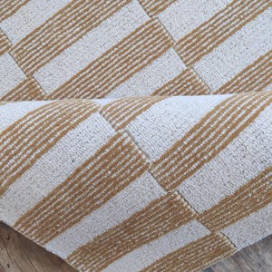 Peut inclure: Gros plan sur un tapis à rayures beiges et marron. Le tapis présente un motif géométrique avec des sections alternées de rayures beiges et marron. Le tapis est partiellement enroulé, révélant le dessous.