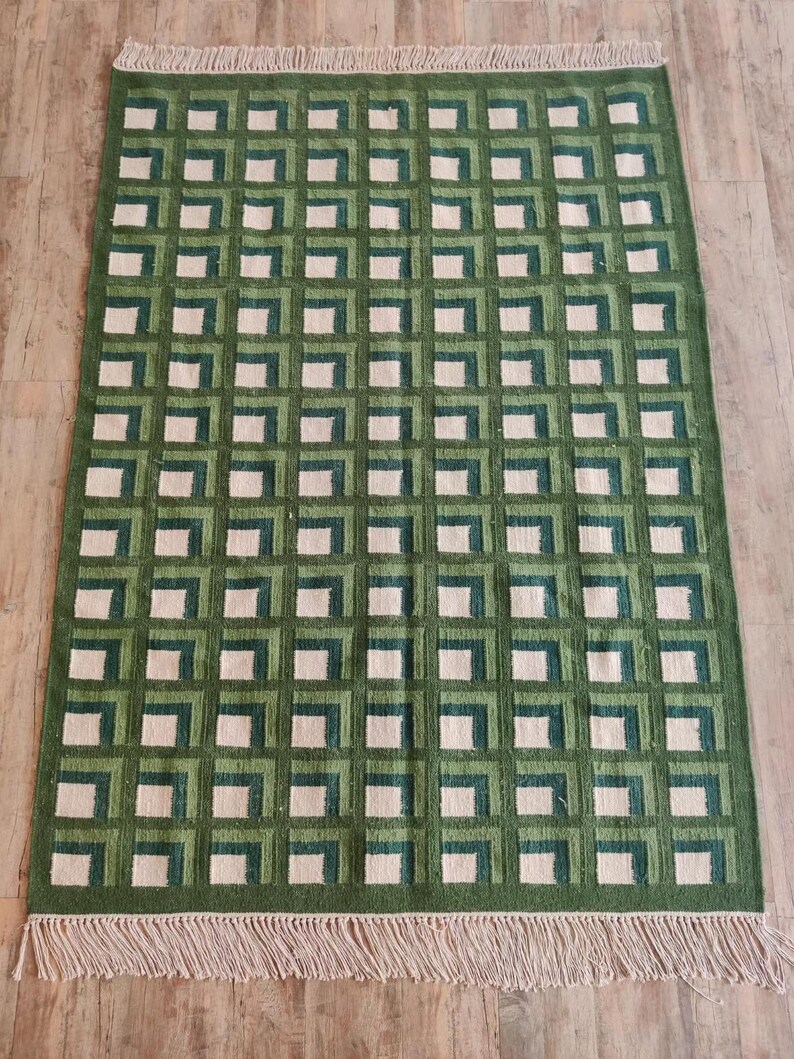 Puede incluir: Una alfombra geom&eacute;trica con estampado verde y blanco con borde de flecos. La alfombra tiene un patr&oacute;n de cuadros con cuadrados de diferentes tonos de verde y blanco.