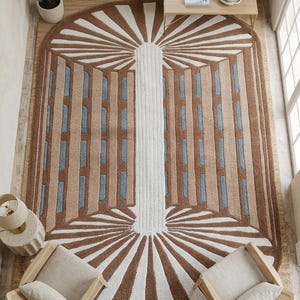 May include: An oval area rug featuring a central white vertical pillar flanked by symmetrical brown and light blue geometric panels. The design includes white sunburst patterns at the top and bottom, creating a structured, layered visual effect.