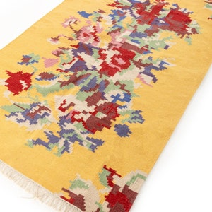 K&ouml;nnte beinhalten: Ein gelber Teppich mit einem bunten geometrischen Muster. Das Muster zeigt die Farben Rot, Blau, Gr&uuml;n und Braun. Der Teppich hat einen Fransenrand.