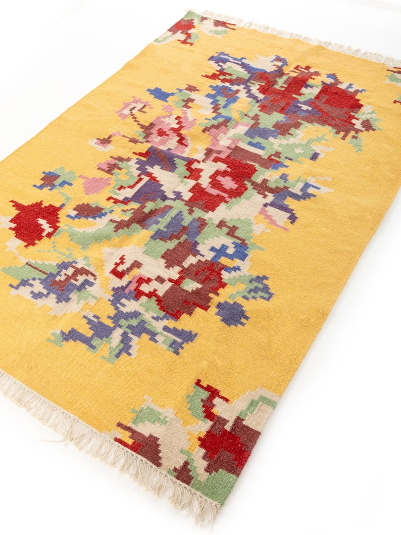 K&ouml;nnte beinhalten: Ein gelber Teppich mit einem bunten geometrischen Muster. Das Muster zeigt die Farben Rot, Blau, Gr&uuml;n und Braun. Der Teppich hat einen Fransenrand.