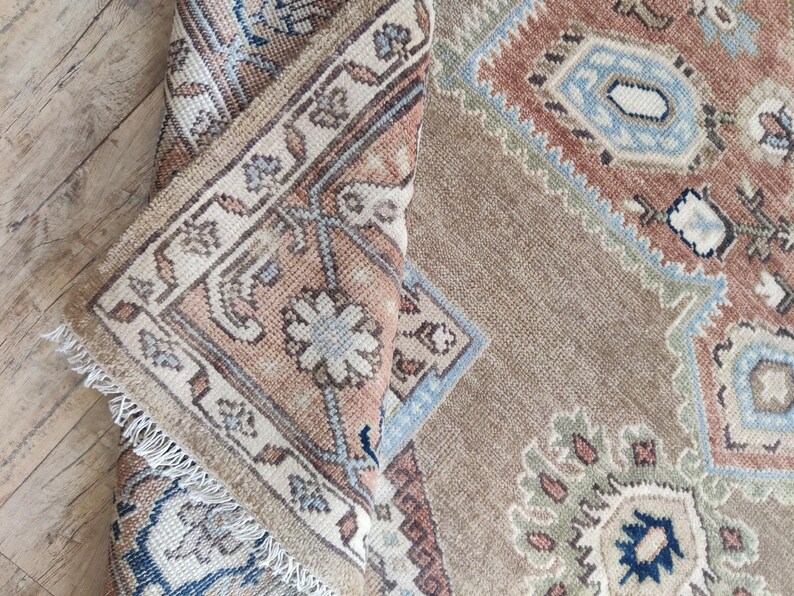 May include: A close-up of a beige and brown rug with a traditional Persian design. The rug has a fringe border and intricate patterns in shades of blue, green, and red.
