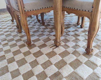 May include: A plush area rug featuring a repeating checkered pattern in alternating squares of cream and light brown. The rug is positioned beneath wooden dining chairs with carved legs and light-colored upholstered seats.