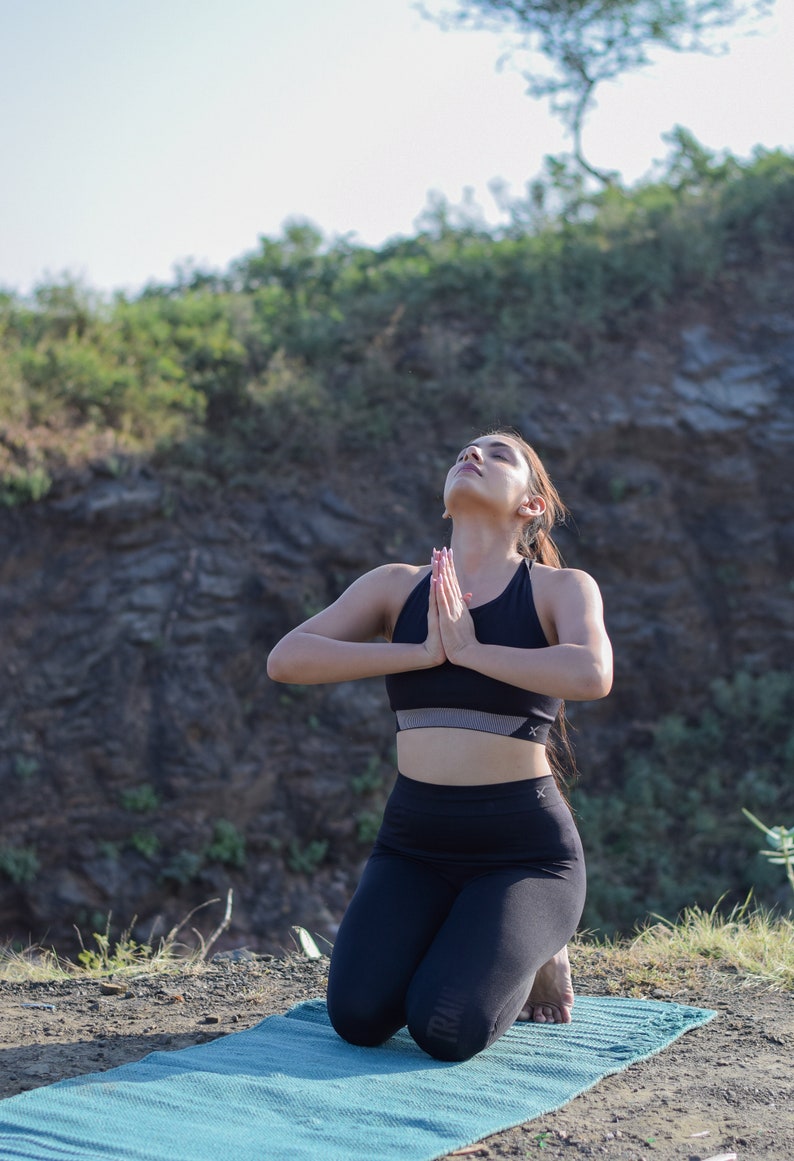 May include: A woman in black athletic wear is kneeling on a blue yoga mat with her hands in prayer position, looking up at the sky. She is practicing yoga outdoors against a backdrop of green foliage and rocky terrain.