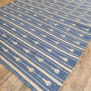Op de afbeelding: Een blauw en wit gestreept tapijt met een subtiel patroon van witte bloemen. Het tapijt is gemaakt van een zacht, geweven materiaal.