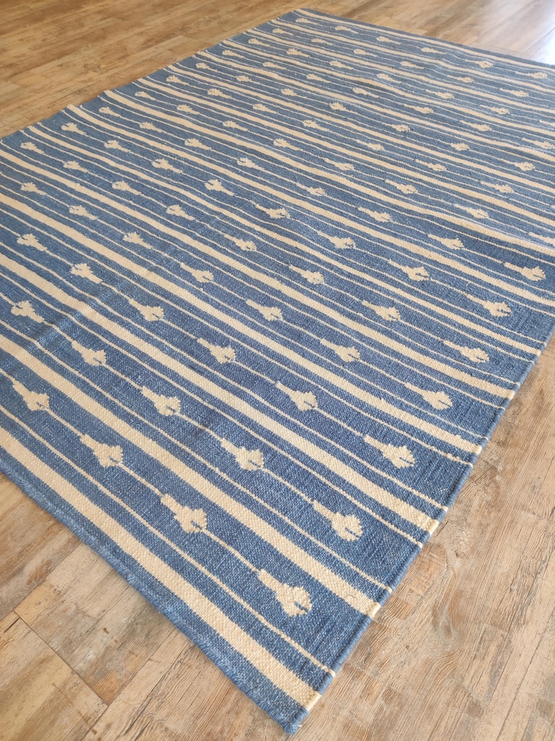 Op de afbeelding: Een blauw en wit gestreept tapijt met een subtiel patroon van witte bloemen. Het tapijt is gemaakt van een zacht, geweven materiaal.