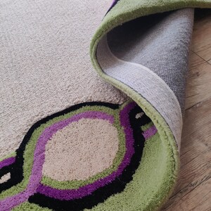 May include: A rolled-up area rug with a beige background and a green, black, and purple circular design. The rug's edge is green, and the design is visible on the rolled-up portion.
