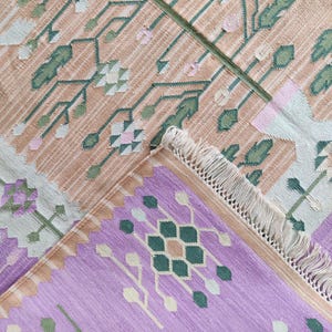 May include: A woven rug with a floral pattern in shades of peach, green, and lavender. The rug features a detailed design with a fringe edge. The design includes geometric and floral elements, creating a textured appearance.