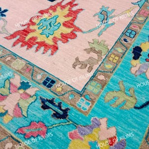 K&ouml;nnte beinhalten: Nahaufnahme eines handgewebten Teppichs mit einem hellrosa Mittelfeld und einem t&uuml;rkisfarbenen Rand. Der Teppich zeigt komplizierte Blumen- und geometrische Muster in Rot, Gelb, Blau und Braun. Der Text "HOUSE OF KILIMS" ist sichtbar.