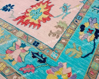 Può includere: Primo piano di un tappeto tessuto a mano con un campo centrale rosa chiaro e un bordo turchese. Il tappeto presenta intricati motivi floreali e geometrici in rosso, giallo, blu e marrone. Il testo "HOUSE OF KILIMS" è visibile.