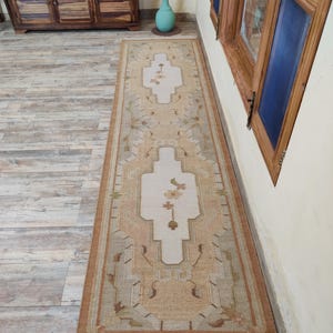 May include: A long, rectangular rug with a cream background and a brown, beige, and green geometric pattern. The rug has a fringe border.
