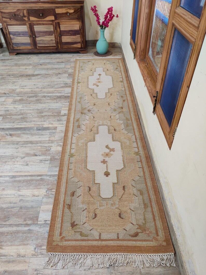 May include: A long, rectangular rug with a cream background and a brown, beige, and green geometric pattern. The rug has a fringe border.