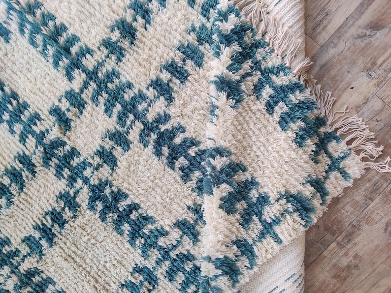 May include: A white and blue patterned rug with a fringe border. The rug has a soft, fluffy texture and is made of wool.